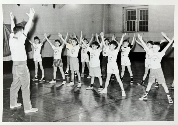 A vintage photo of a YMCA gym class with boys in athletic attire performing exercises under the supervision of an instructor.